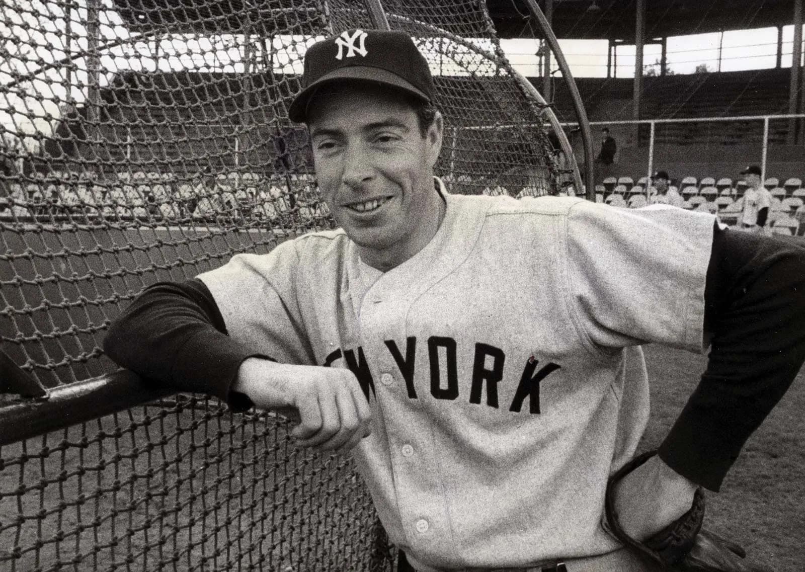 Black and white photo of legendary baseball record-setter Joe DiMaggio in uniform, leaning against a fence at a baseball field.